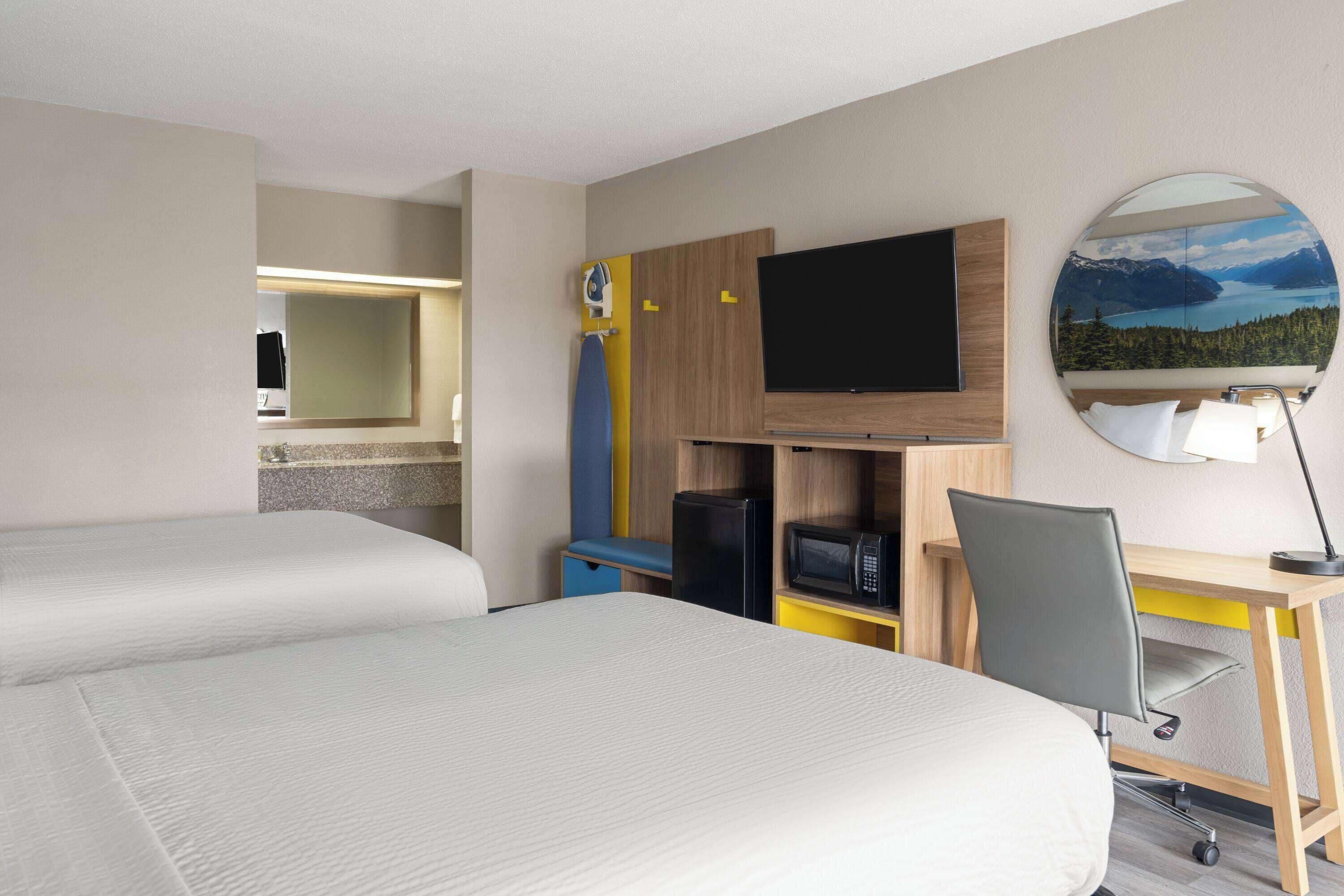 Habitación de hotel con dos camas, un escritorio con silla, televisión, espejo y vistas a las montañas.