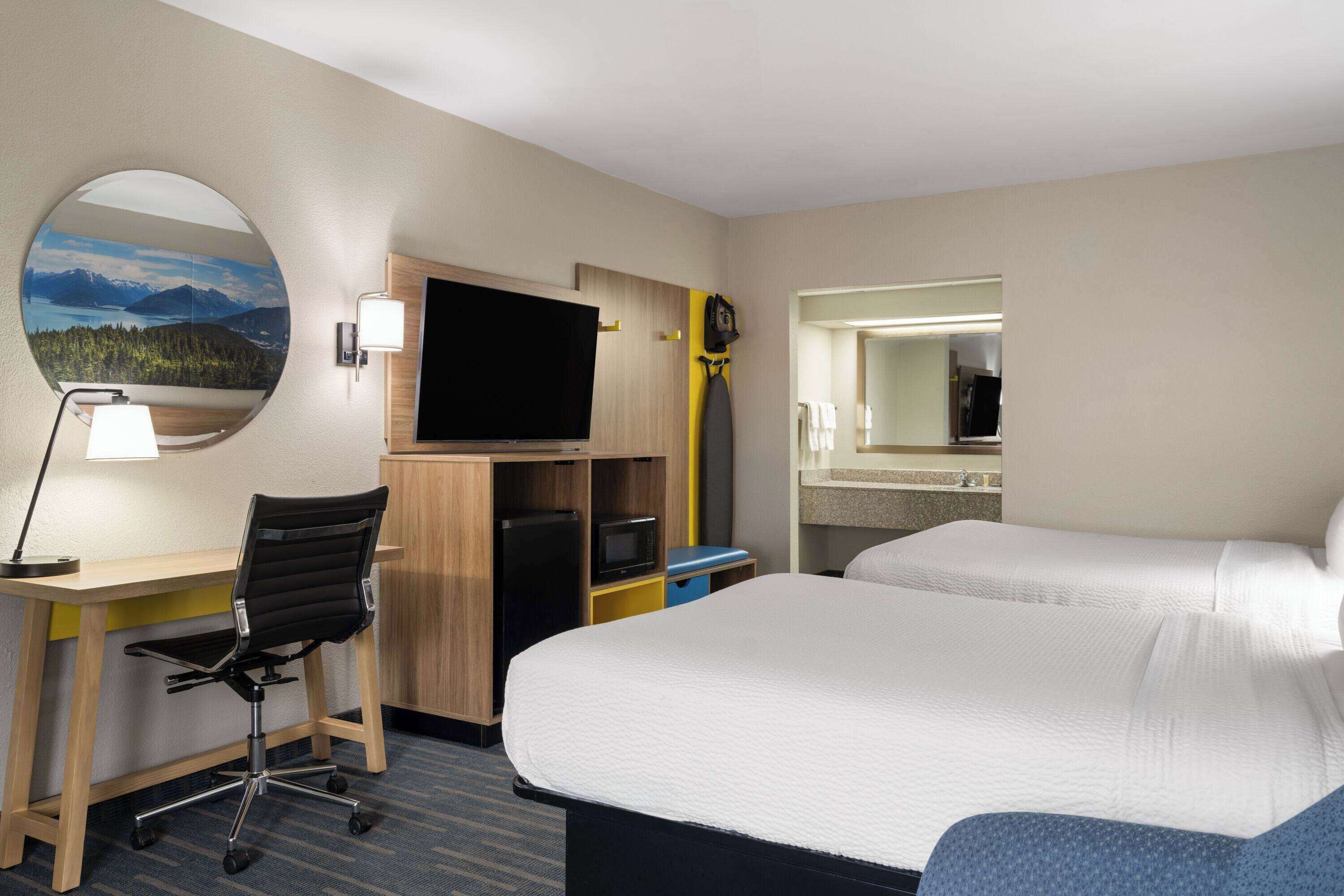 A hotel room with a bed, a desk with a chair, a television, a mirror, and a view of mountains.