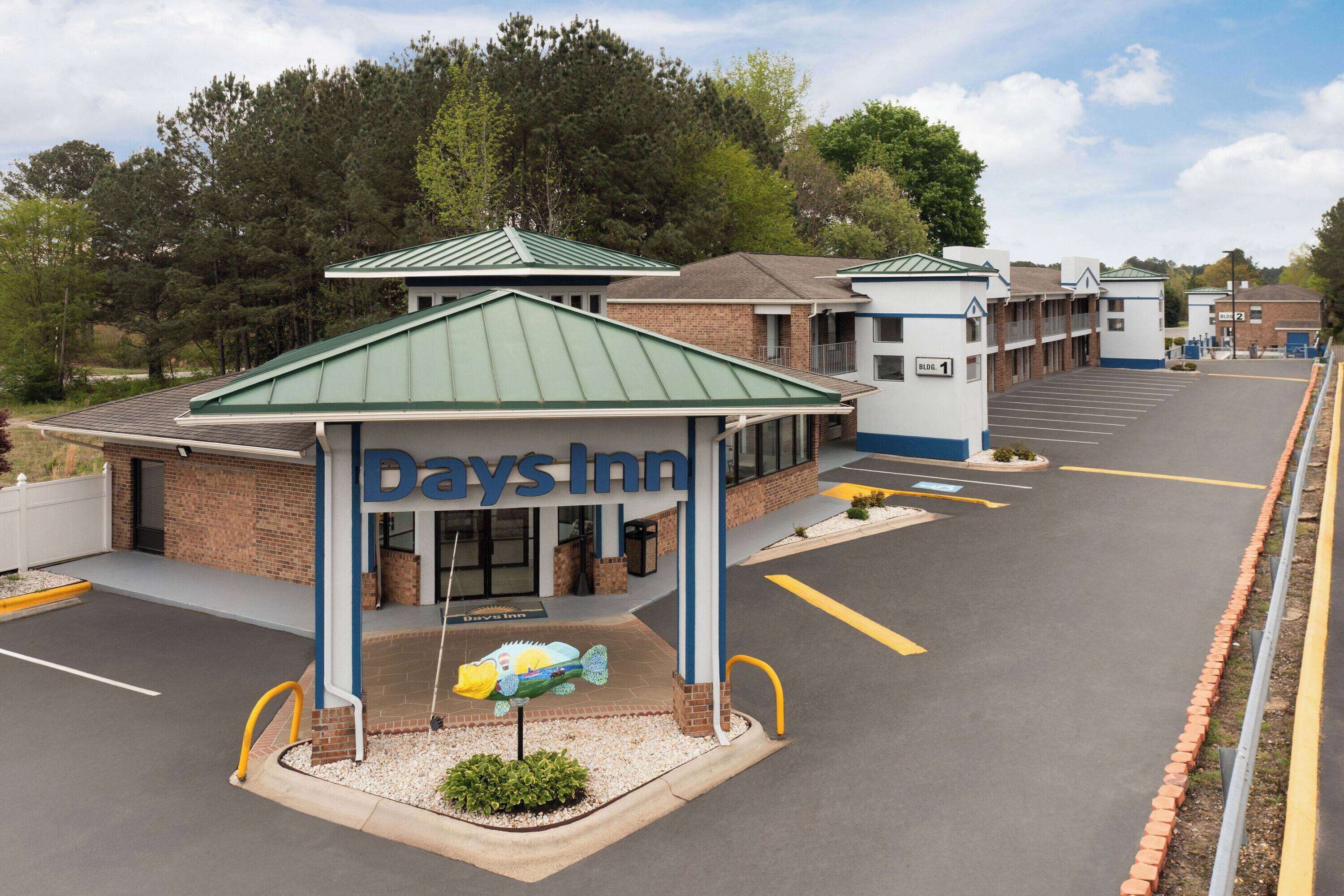 A Days Inn with a green roof, a parking lot, and a small garden area with a decorative fish sculpture.