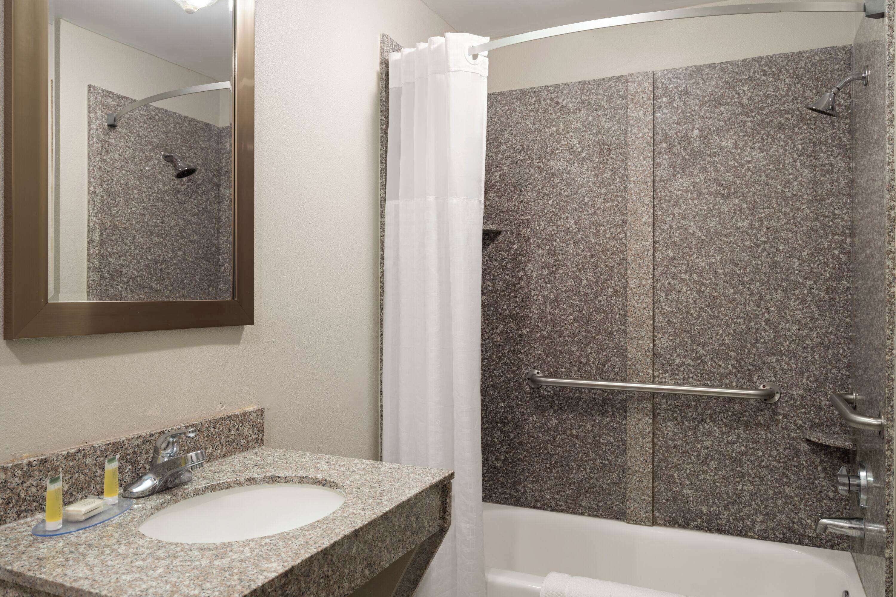 Baño con lavamanos de encimera de granito, espejo, ducha con cabezal de ducha tipo lluvia y barra de apoyo.