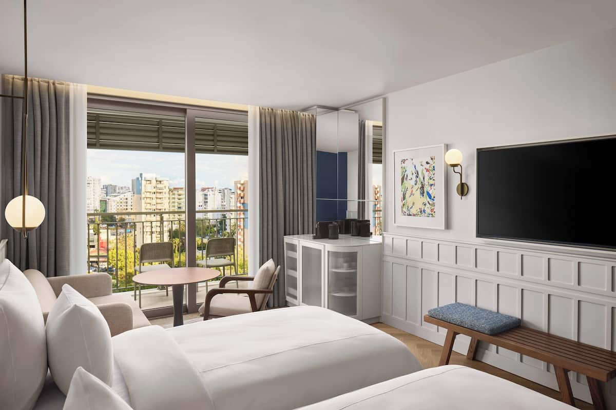 A modern hotel room with a large bed, a desk, a TV, and a balcony with a city view.