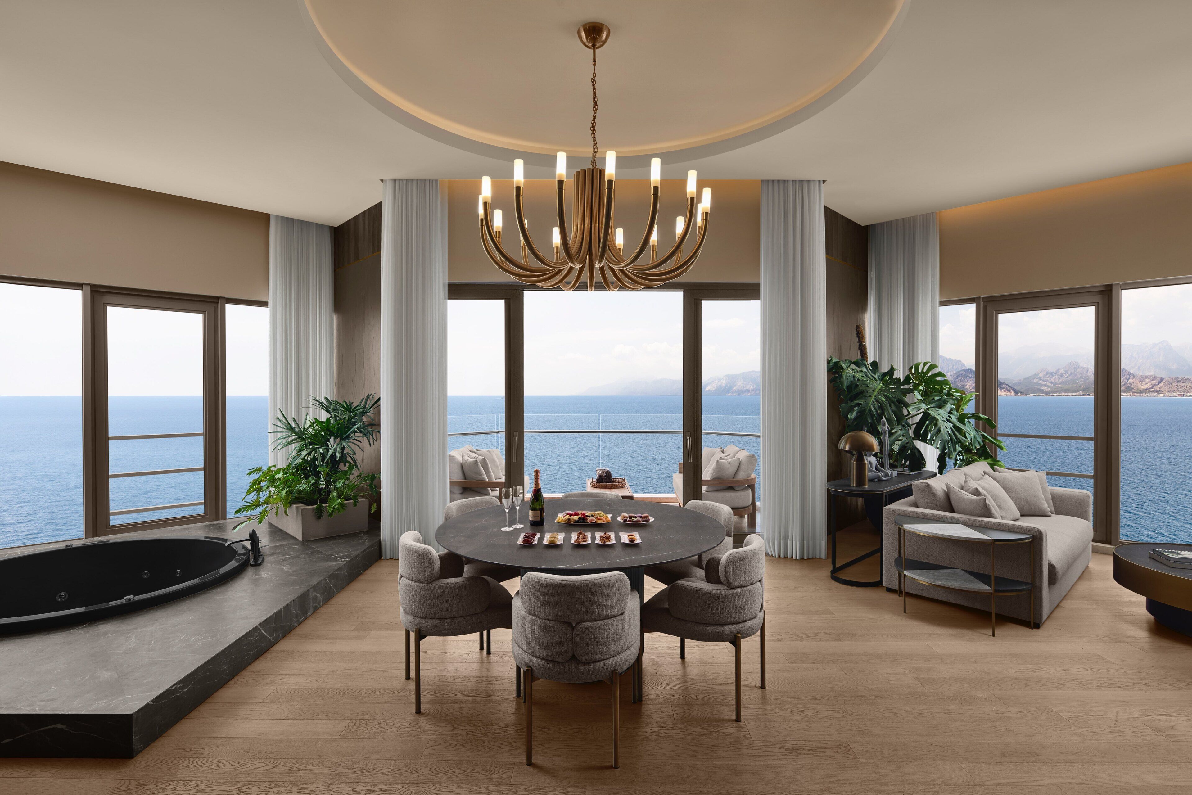 A spacious room with a round dining table, comfortable seating, a large window overlooking the sea, and a modern chandelier.