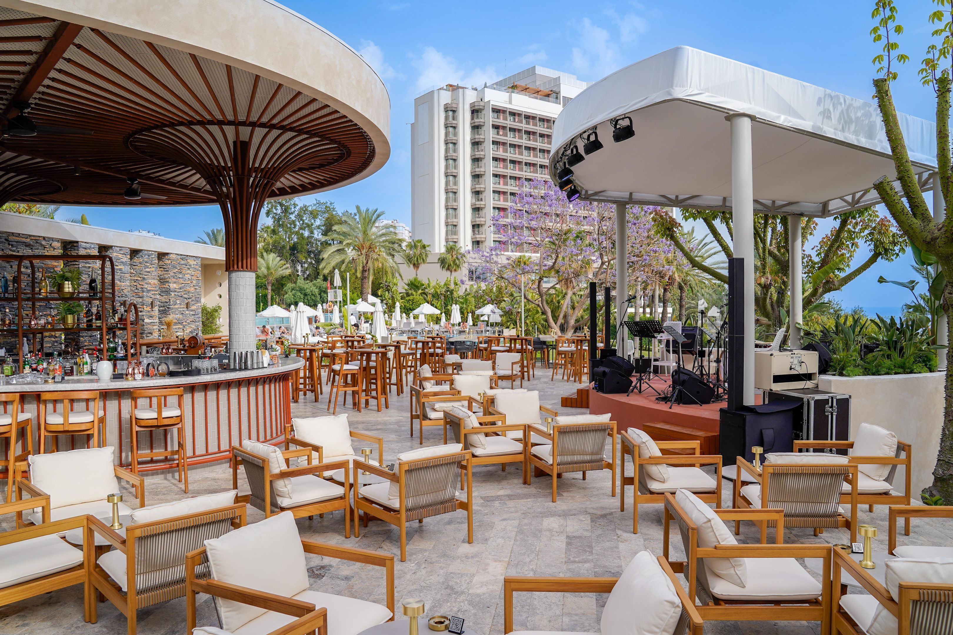 An outdoor bar area with wooden chairs and tables, a stage with musical equipment, and a view of a high-rise building in the background.