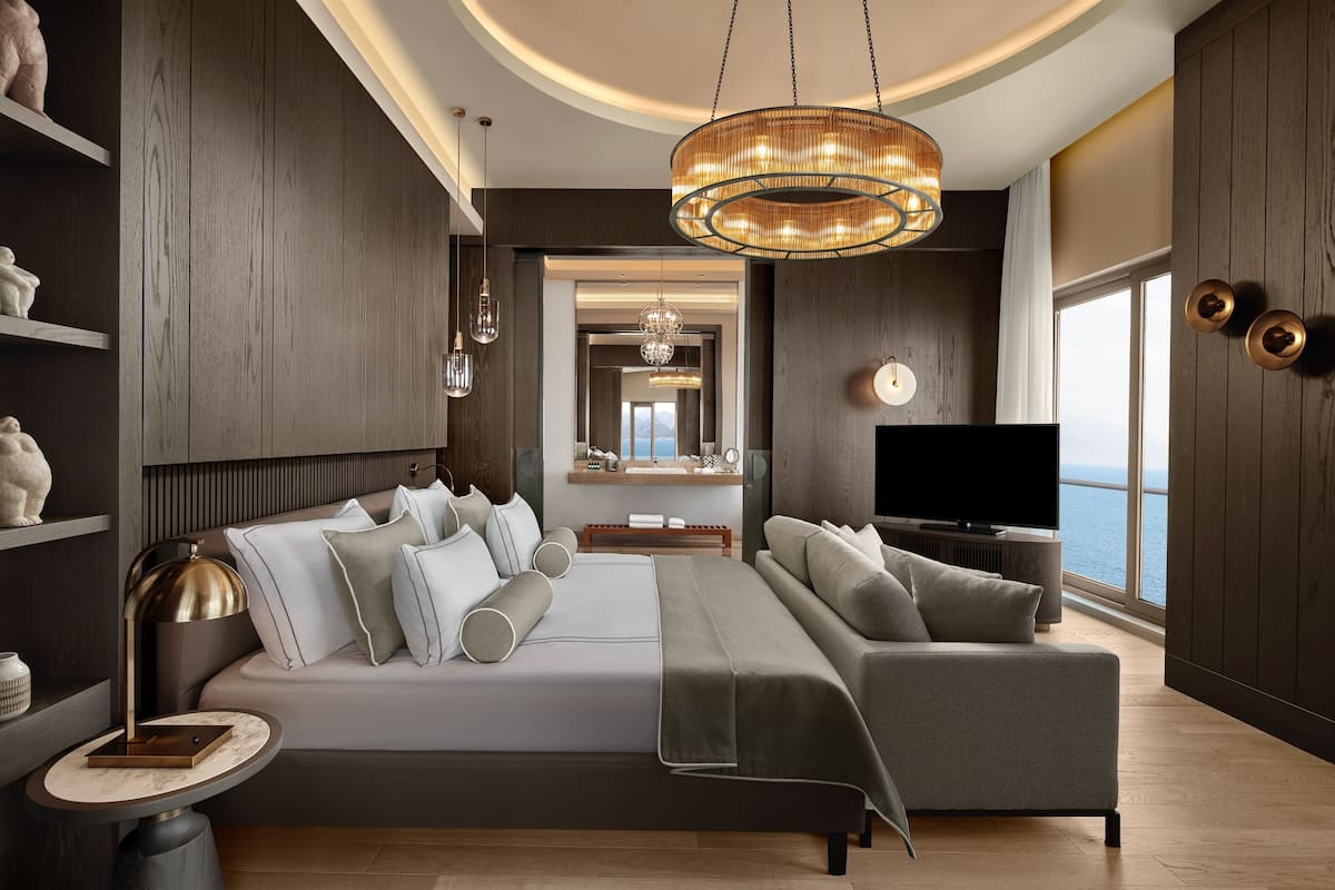 A modern bedroom with a large bed, a sofa, a TV, and a view of the sea.