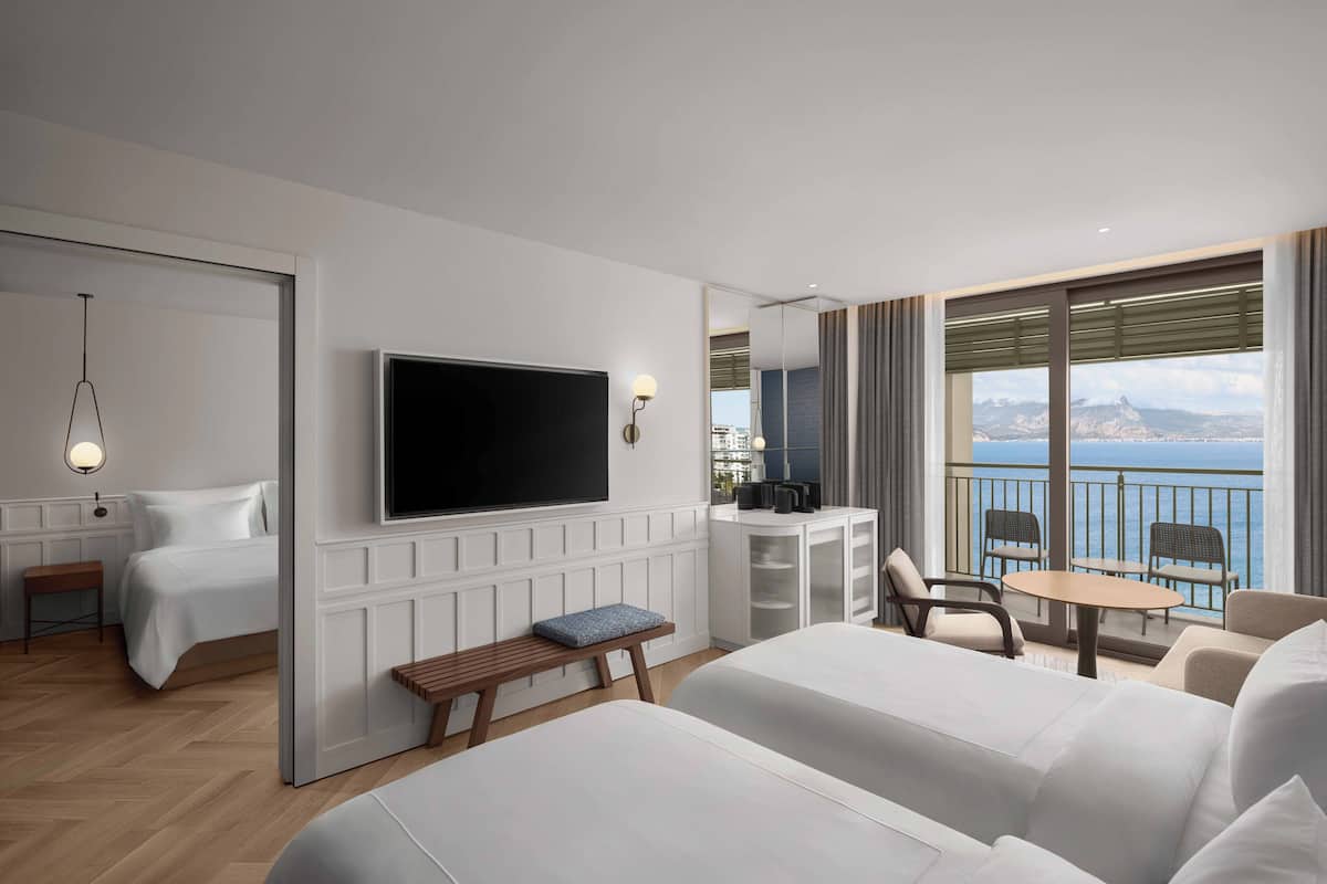 A modern hotel room with a large bed, a TV, a balcony with seating, and a view of the sea.