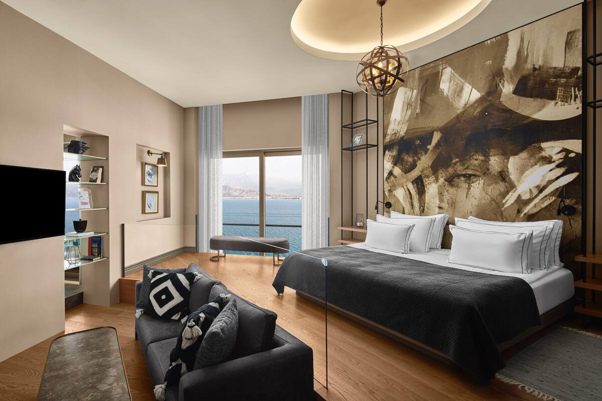 A modern hotel room with a large bed, a sofa, a TV, and a view of the sea.