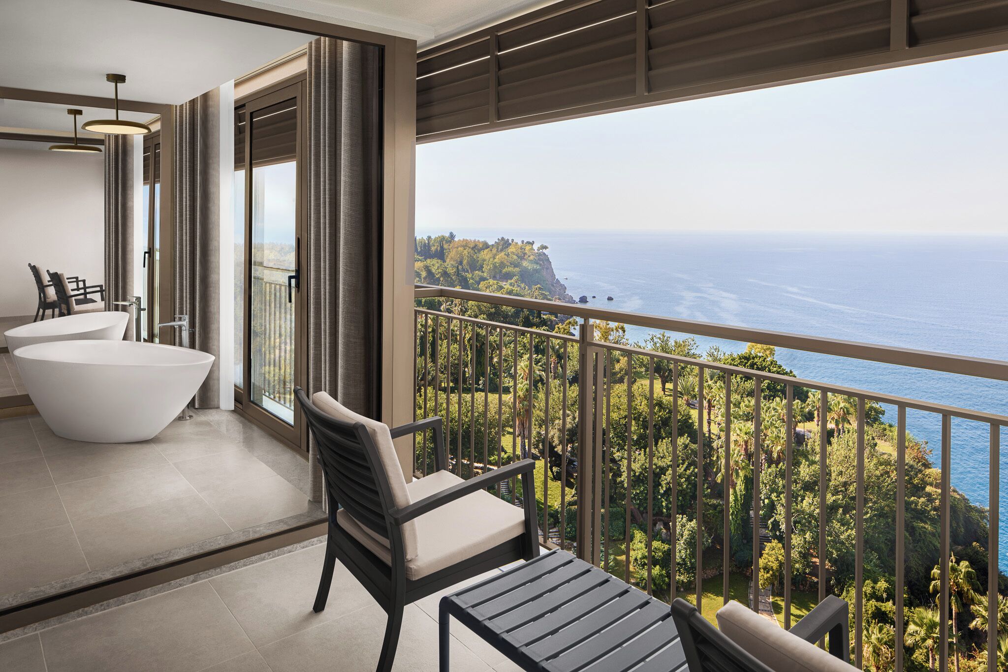 A balcony with a view of the ocean and greenery, a white bathtub, and a seating area with chairs.