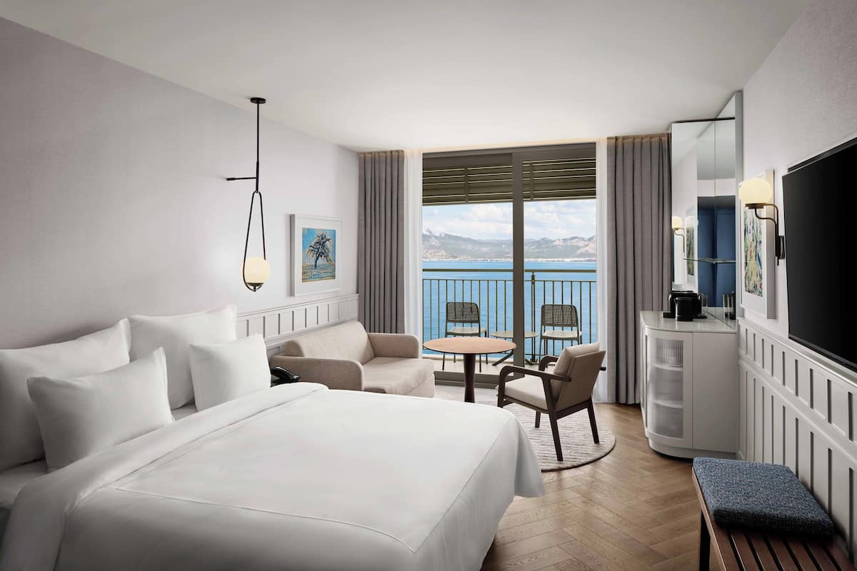A hotel room with a large bed, a desk, a chair, a small table, and a balcony with a view of the sea.