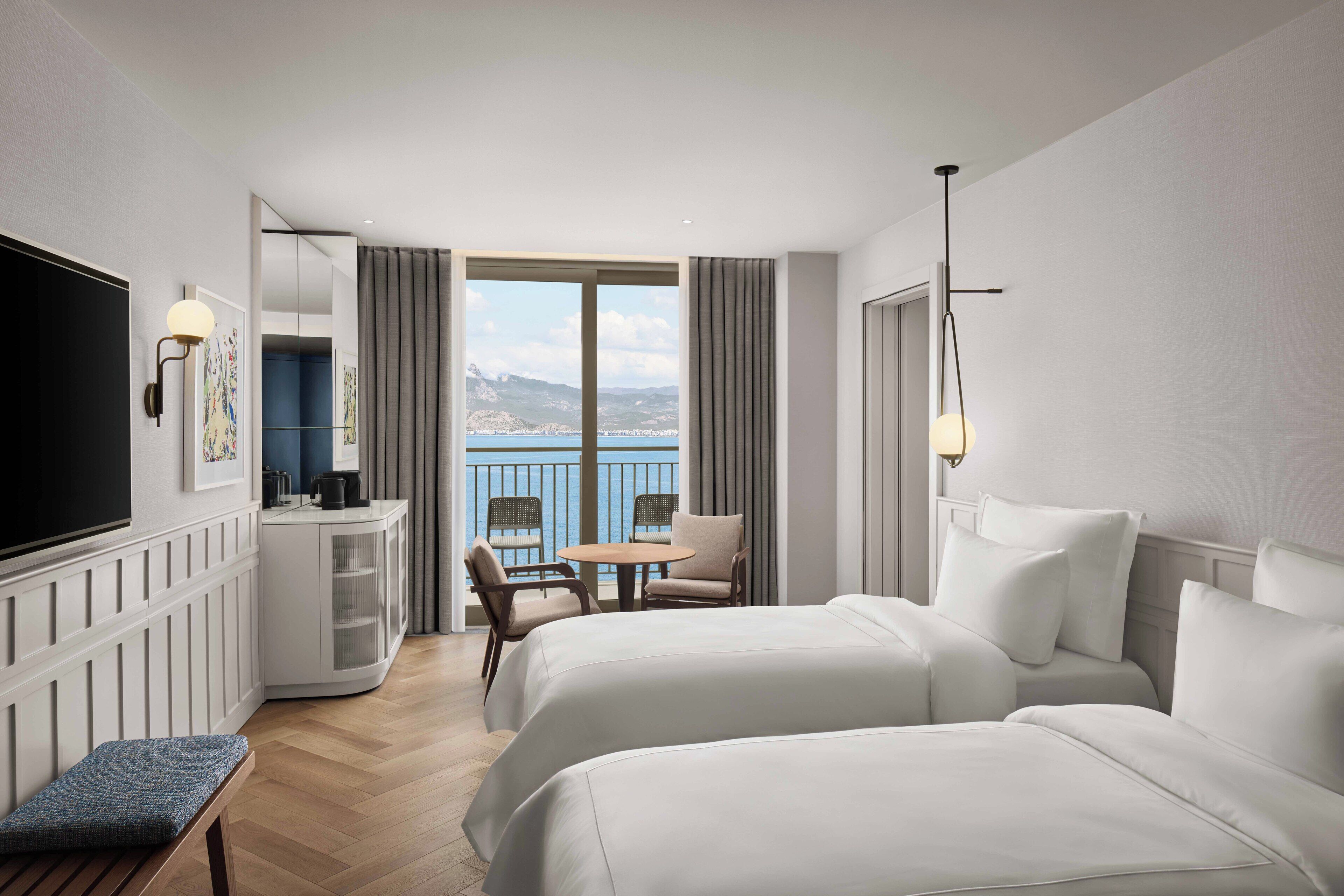 A hotel room with two beds, a TV, a desk with a chair, a balcony with a view, and a small table.