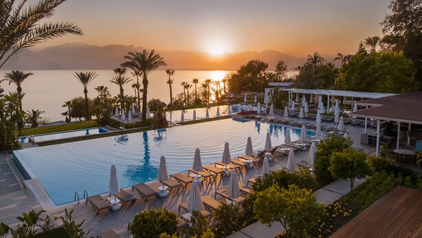 A resort pool with lounge chairs and umbrellas, surrounded by palm trees and a view of the sunset over the water. - Akra Antalya (Antalya)