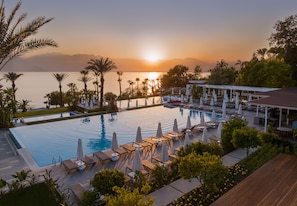 A resort pool with lounge chairs and umbrellas, surrounded by palm trees and a view of the sunset over the water. - Akra Antalya (Antalya)
