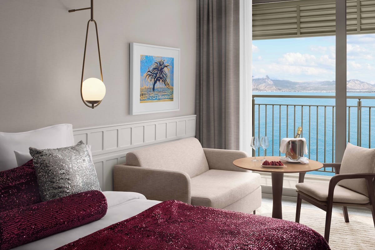 A hotel room with a bed, a sofa, a small table with a bottle and glasses, and a view of the sea.