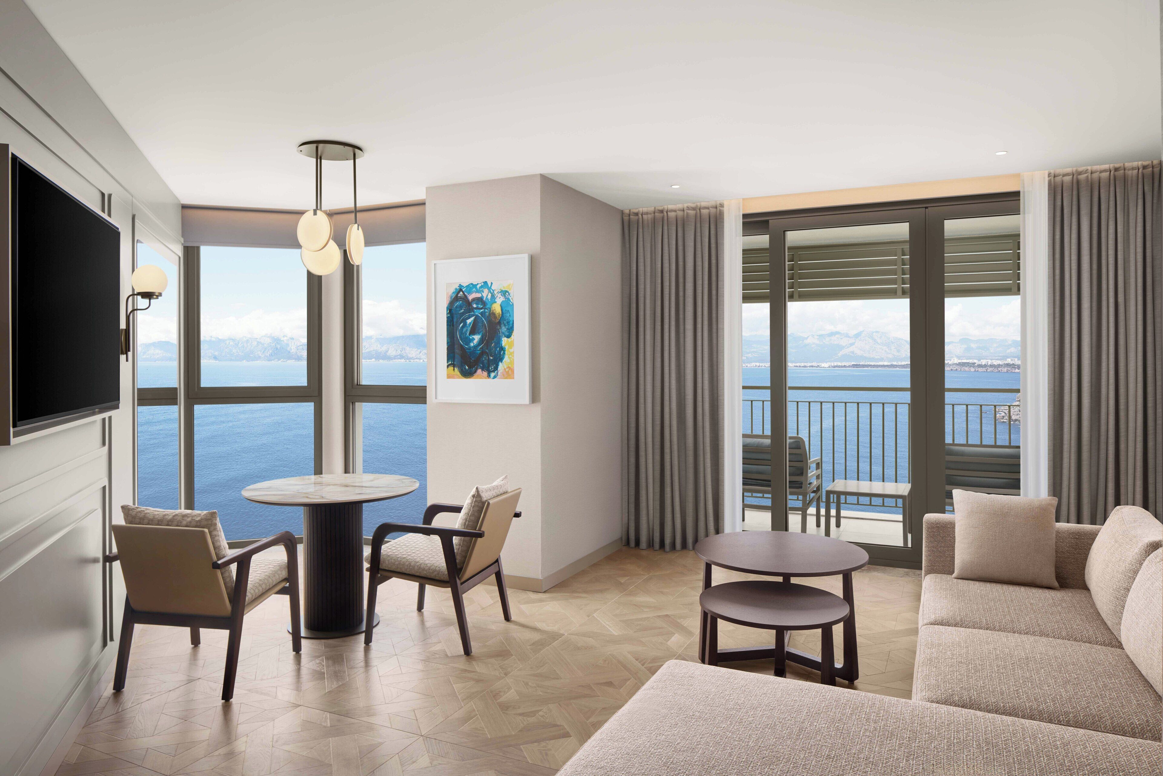 A modern hotel room with a large window offering a scenic view of the sea and mountains. There is a comfortable seating area with a sofa and chairs, a small round table, and a flat-screen TV mounted on the wall.
