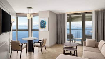 A modern hotel room with a large window offering a scenic view of the sea and mountains. There is a comfortable seating area with a sofa and chairs, a small round table, and a flat-screen TV mounted on the wall.