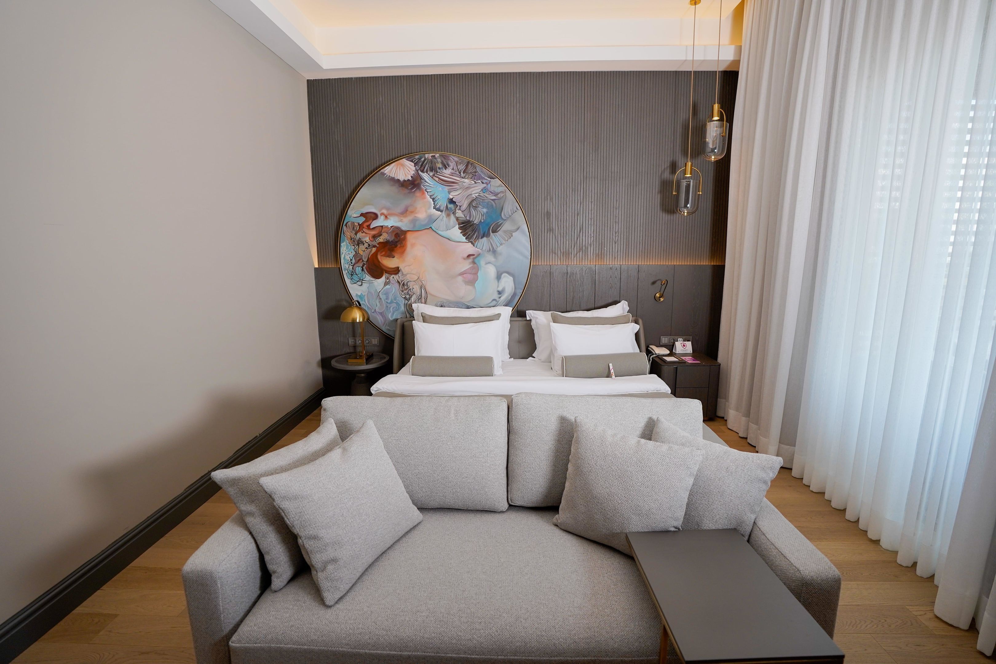 A modern hotel room with a large bed, a grey sofa, and a round painting on the wall.