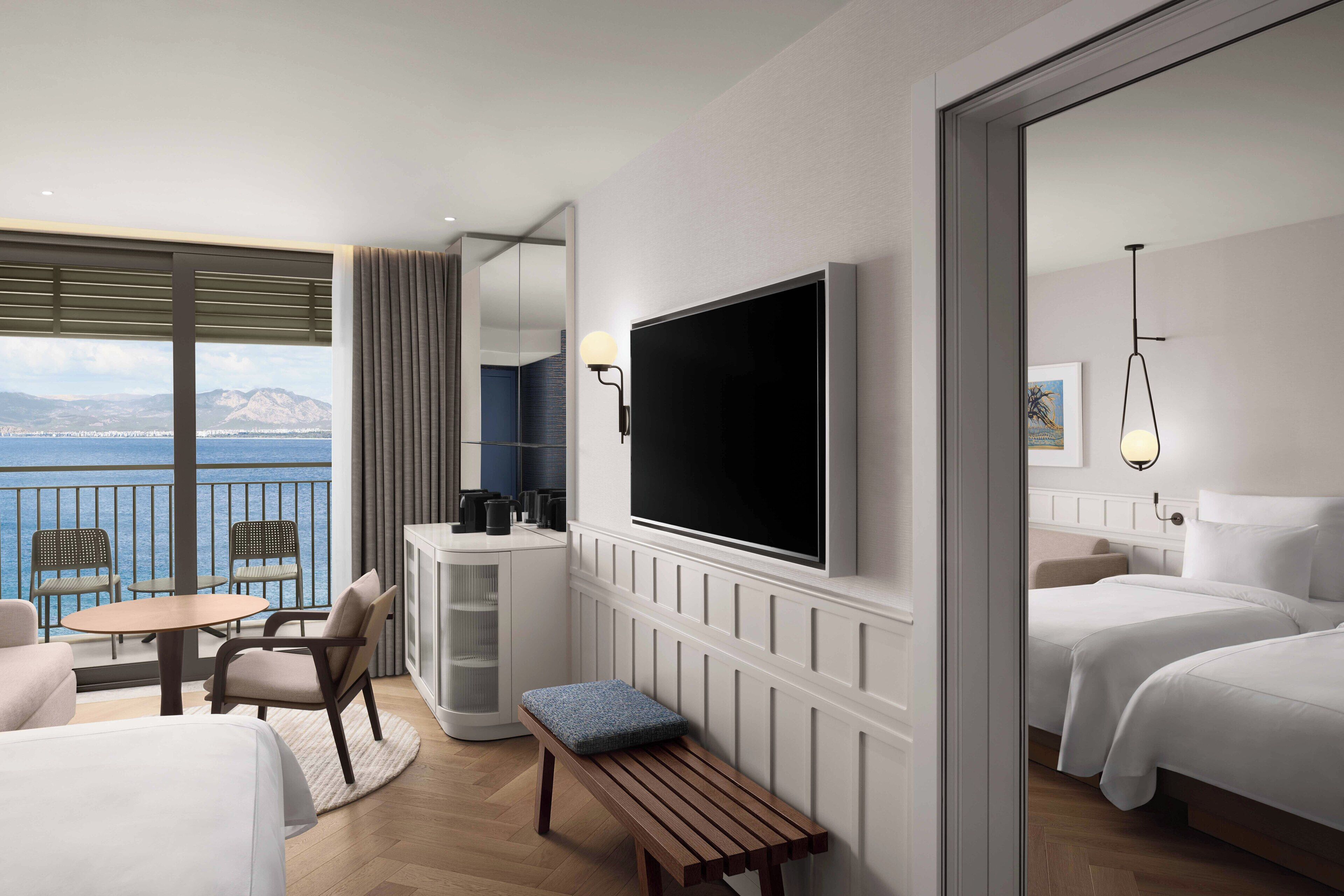 A hotel room with a balcony, a TV, a bed, a bench, and a seating area with chairs and a table.