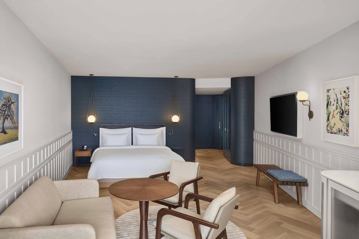 A hotel room with a large bed, a round wooden table, two armchairs, a bench, a TV, and artwork on the walls.