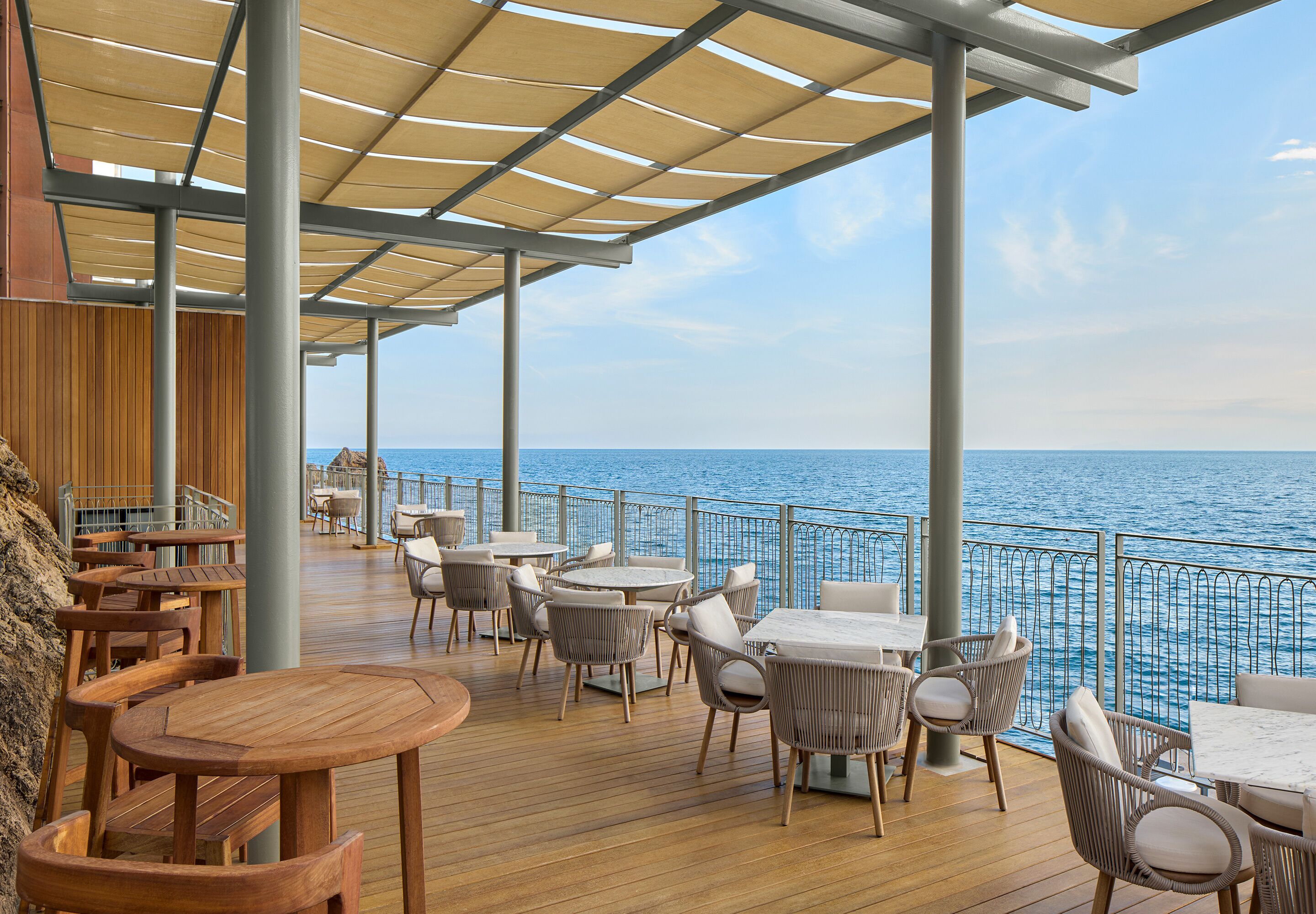 Una terraza con mesas y sillas de madera, barandilla y vista al mar.