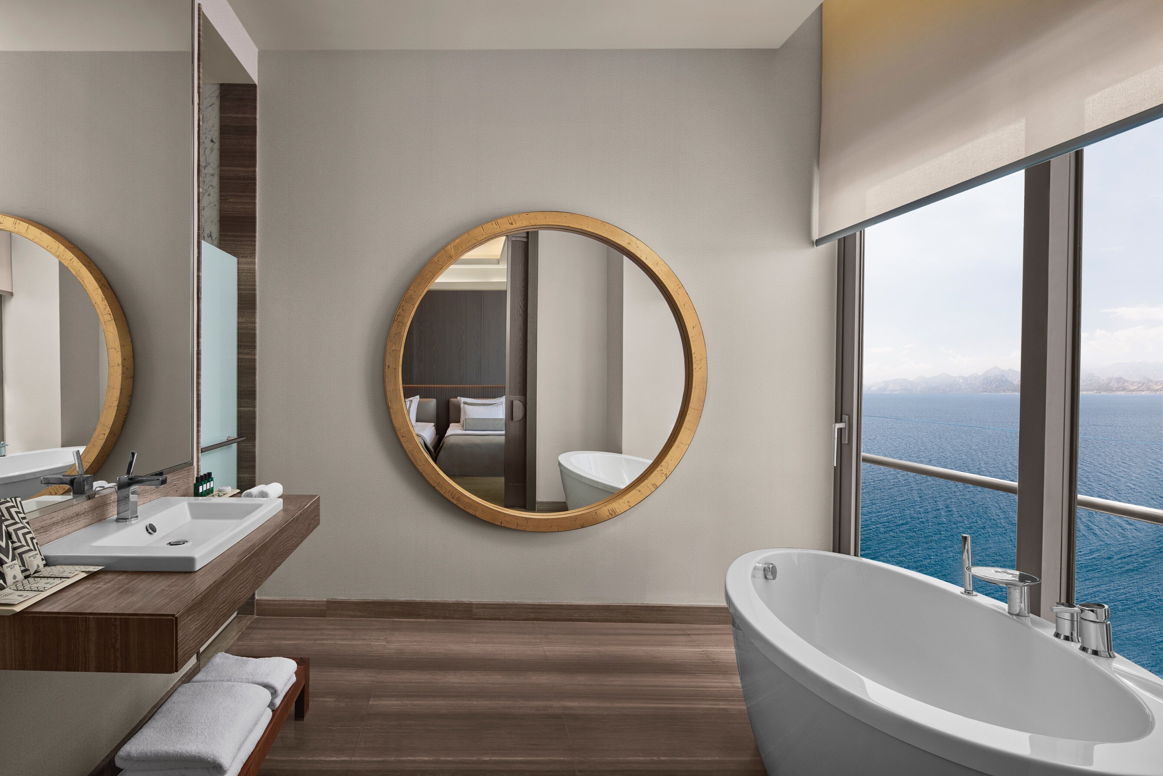Ein modernes Badezimmer mit einer großen Badewanne, einem Waschtisch und einem großen Spiegel. Durch ein großes Fenster ist der Blick aufs Meer.