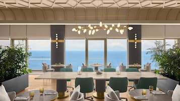 A modern dining area with a large table and chairs, large windows with a view of the sea, and a unique ceiling design.