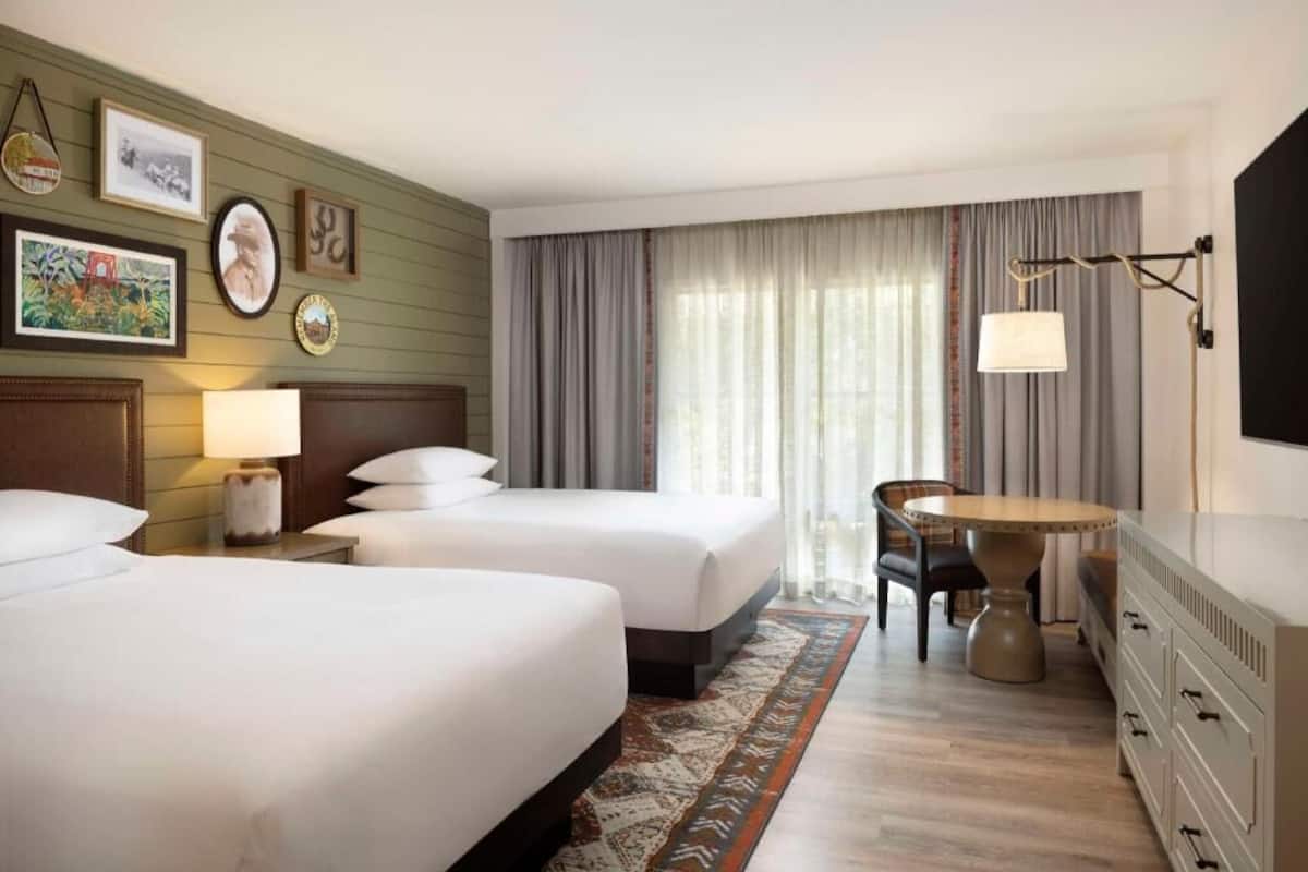 Une chambre d’hôtel avec deux lits, un bureau avec une chaise, une télévision et une fenêtre avec des rideaux.