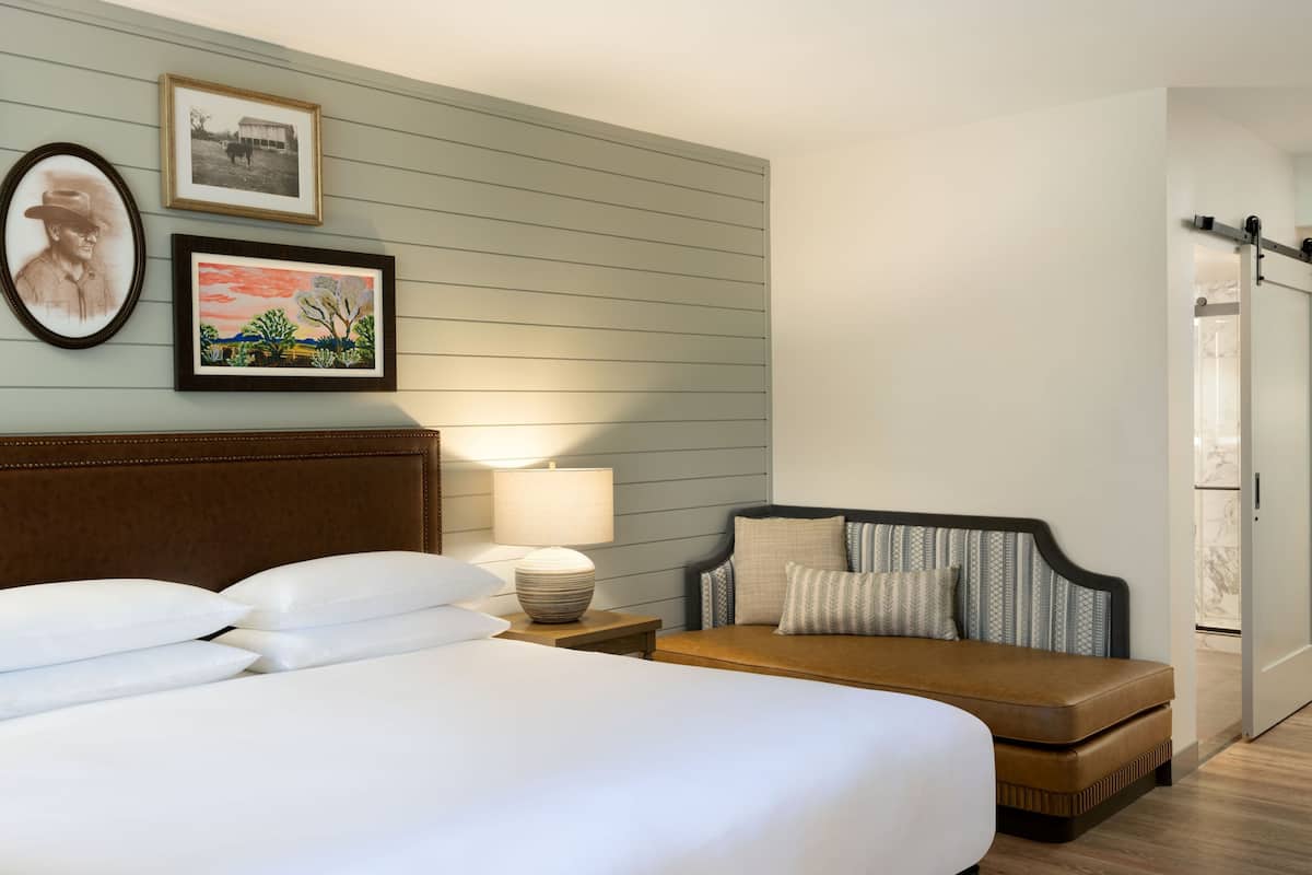 Une chambre d’hôtel avec un grand lit, une table de chevet avec une lampe, un canapé et deux tableaux encadrés au mur.