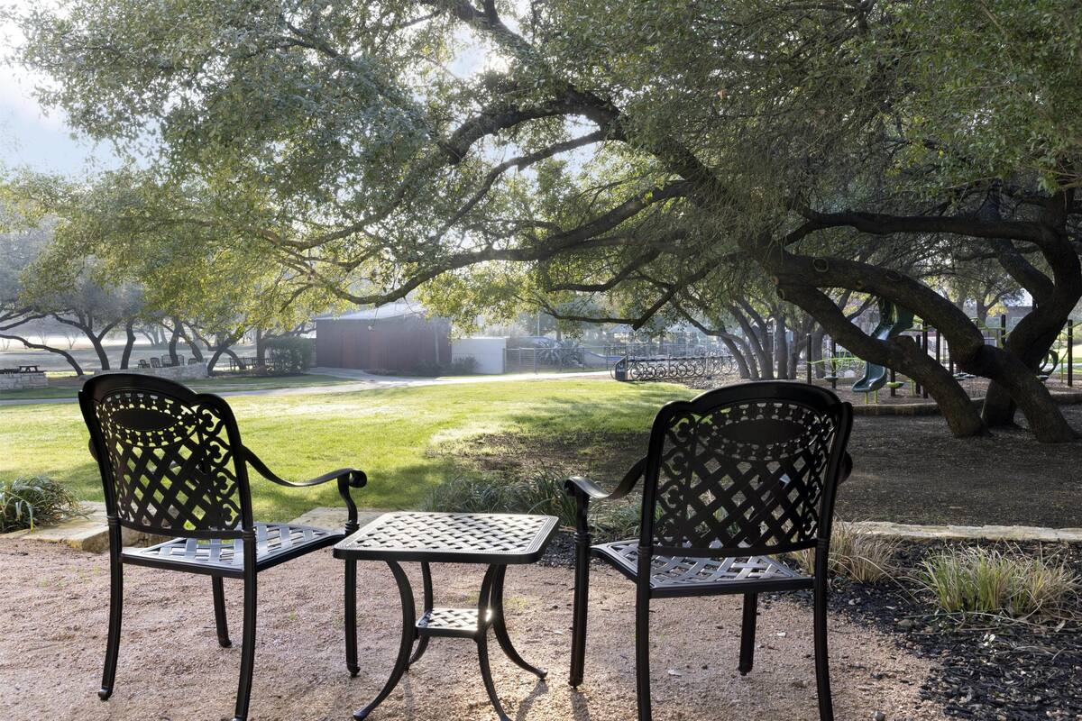 Deux chaises en métal et une table, installées en plein air dans un parc, avec un grand arbre et un bâtiment en arrière-plan.