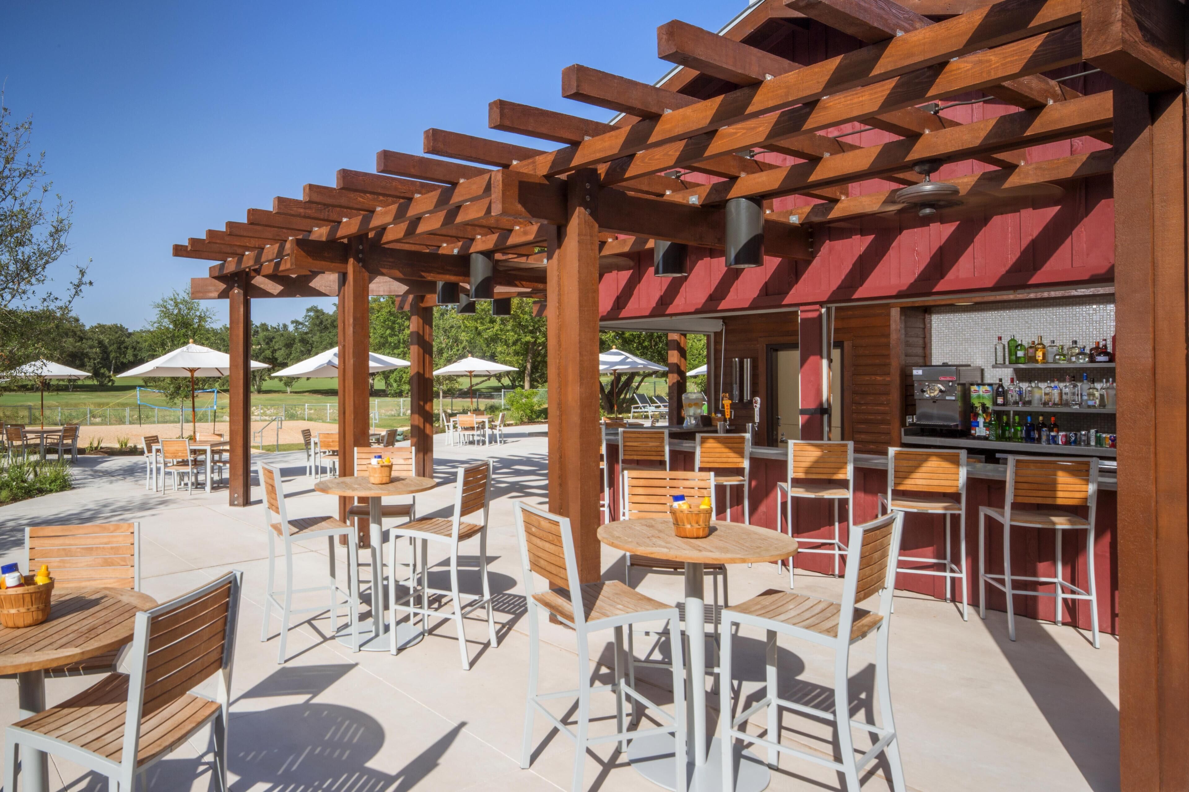 An outdoor patio with wooden tables and chairs, a bar area with bottles, and umbrellas.