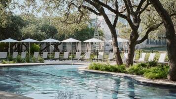 Una piscina con sillones de descanso y sombrillas, rodeada de árboles y un edificio de piedra al fondo.