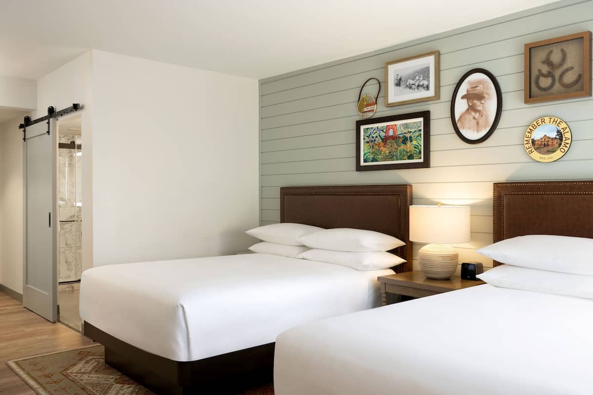 Une chambre d’hôtel avec deux lits, une table de chevet avec une lampe, un cadre photo fixé au mur et un espace douche.