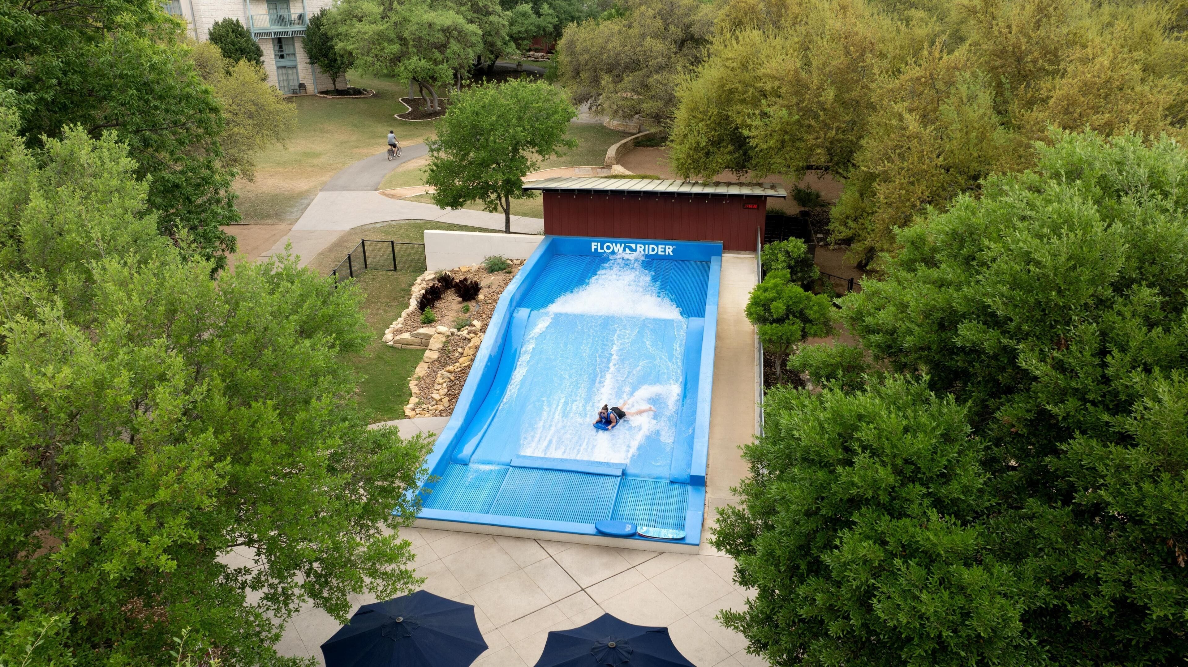 A water slide with a person at the bottom, surrounded by trees and a building with a sign 'FLOW RIDER'.