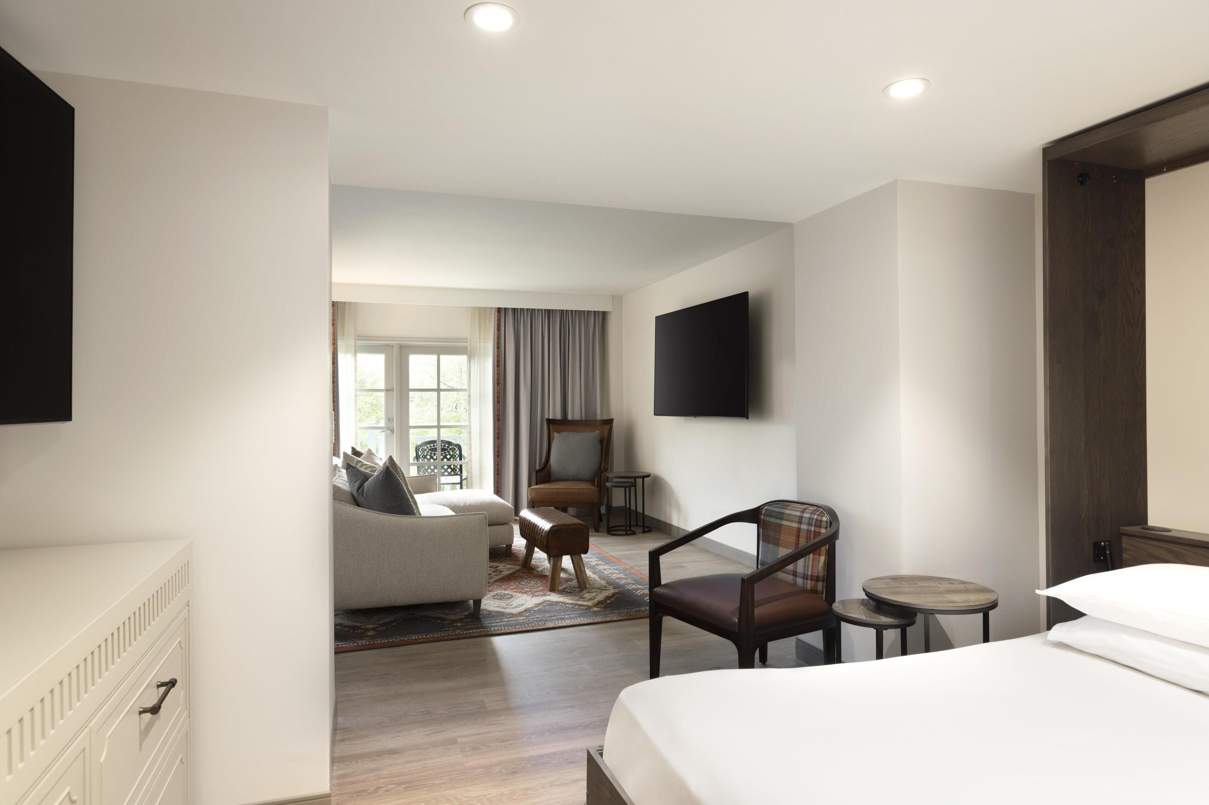 Habitación de hotel con cama, sofá, sillón, una mesita, televisor y ventana con cortinas.
