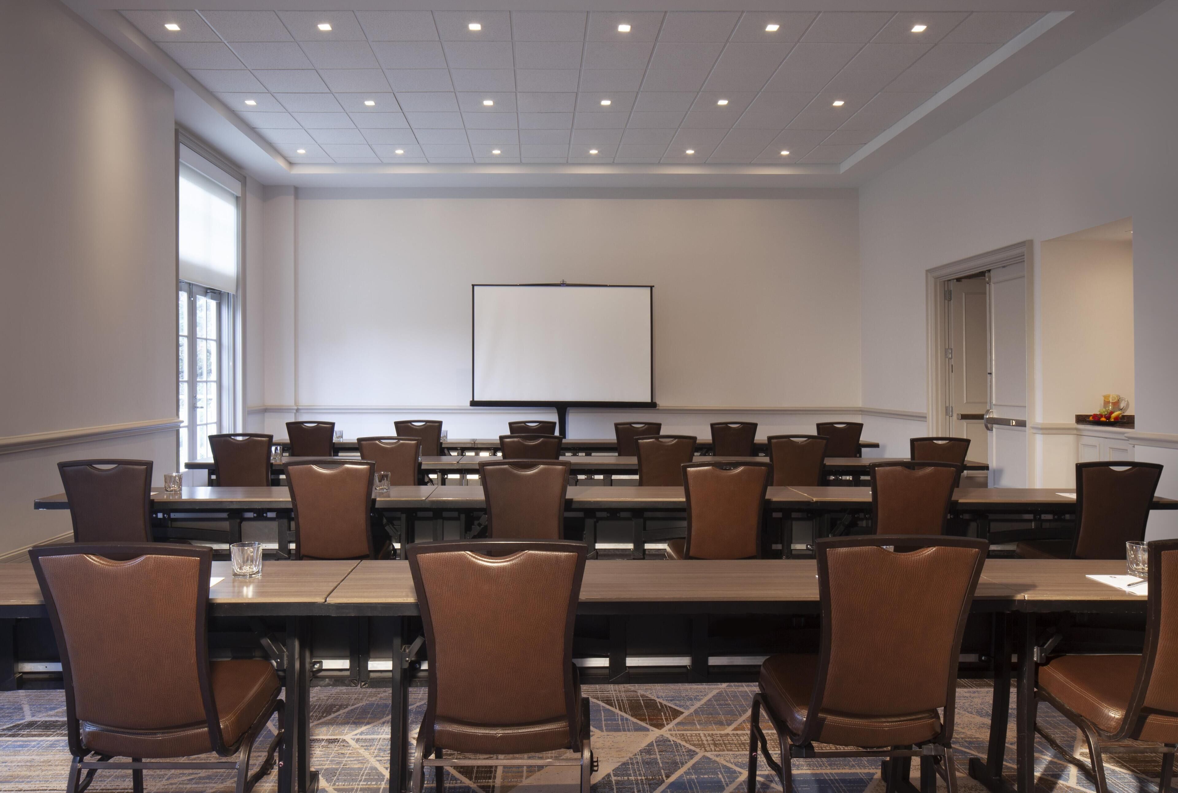 A conference room with multiple tables and chairs, a projector screen, and a large window.