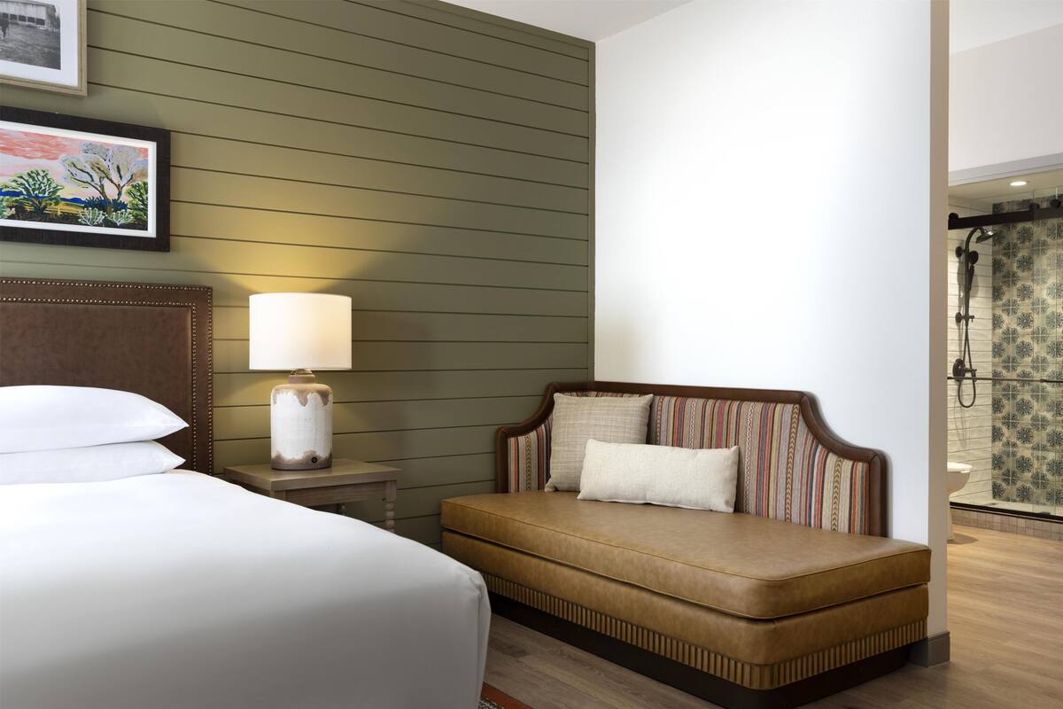 Une chambre d’hôtel comprenant un lit, une table de chevet avec une lampe, un canapé agrémenté de coussins à motifs et un espace douche avec des carreaux à motifs.