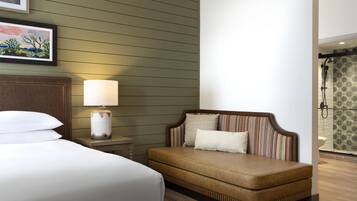 A hotel room with a bed, a bedside table with a lamp, a sofa with patterned cushions, and a shower area with patterned tiles.