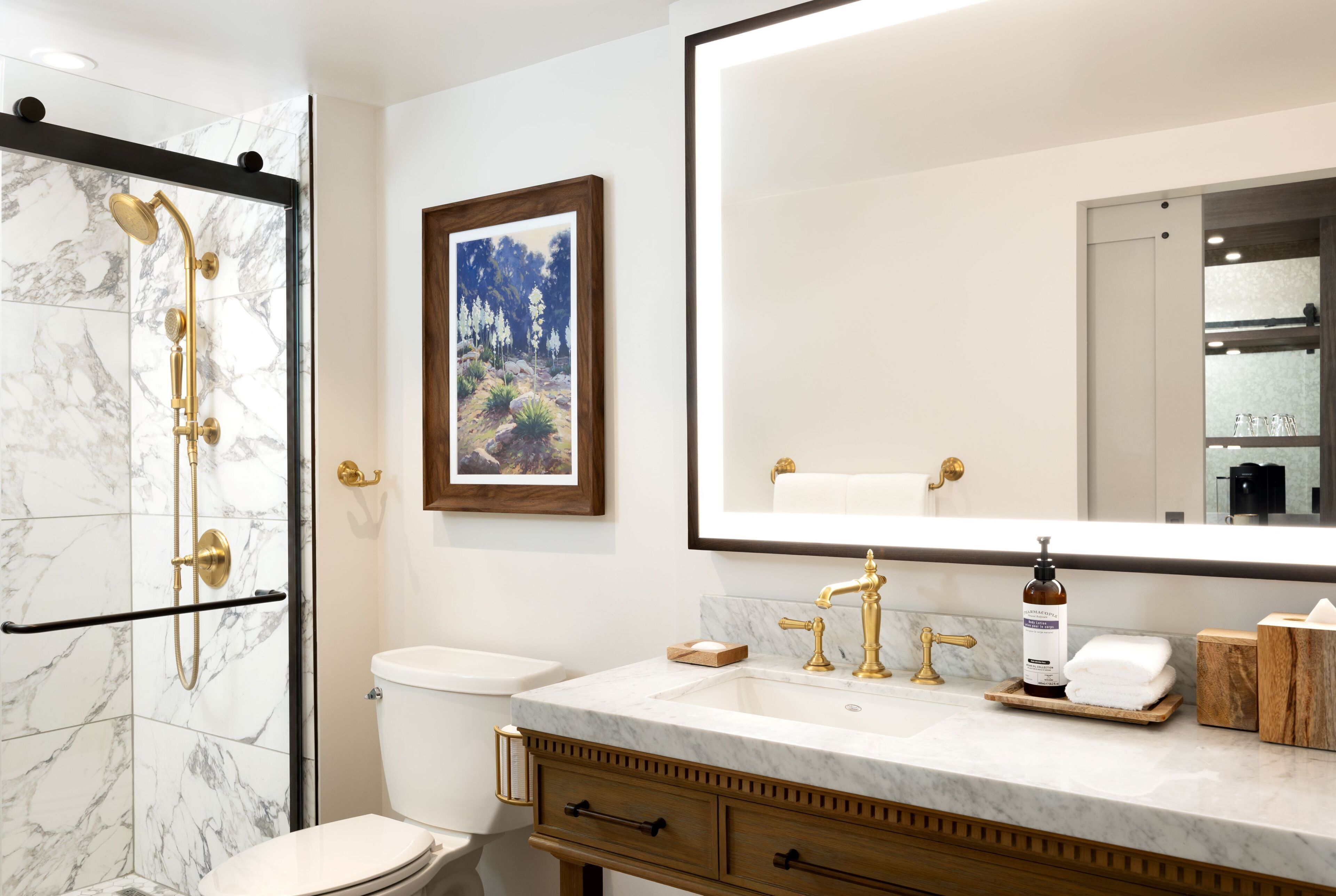 Un baño moderno con ducha de mármol, inodoro, lavamanos con detalles dorados y un cuadro enmarcado en la pared.