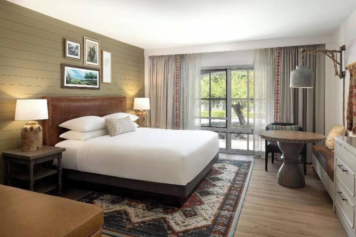 Une chambre d’hôtel avec un grand lit, deux tables de chevet avec des lampes, une petite table avec des chaises, une commode et une vue sur la verdure par la fenêtre.