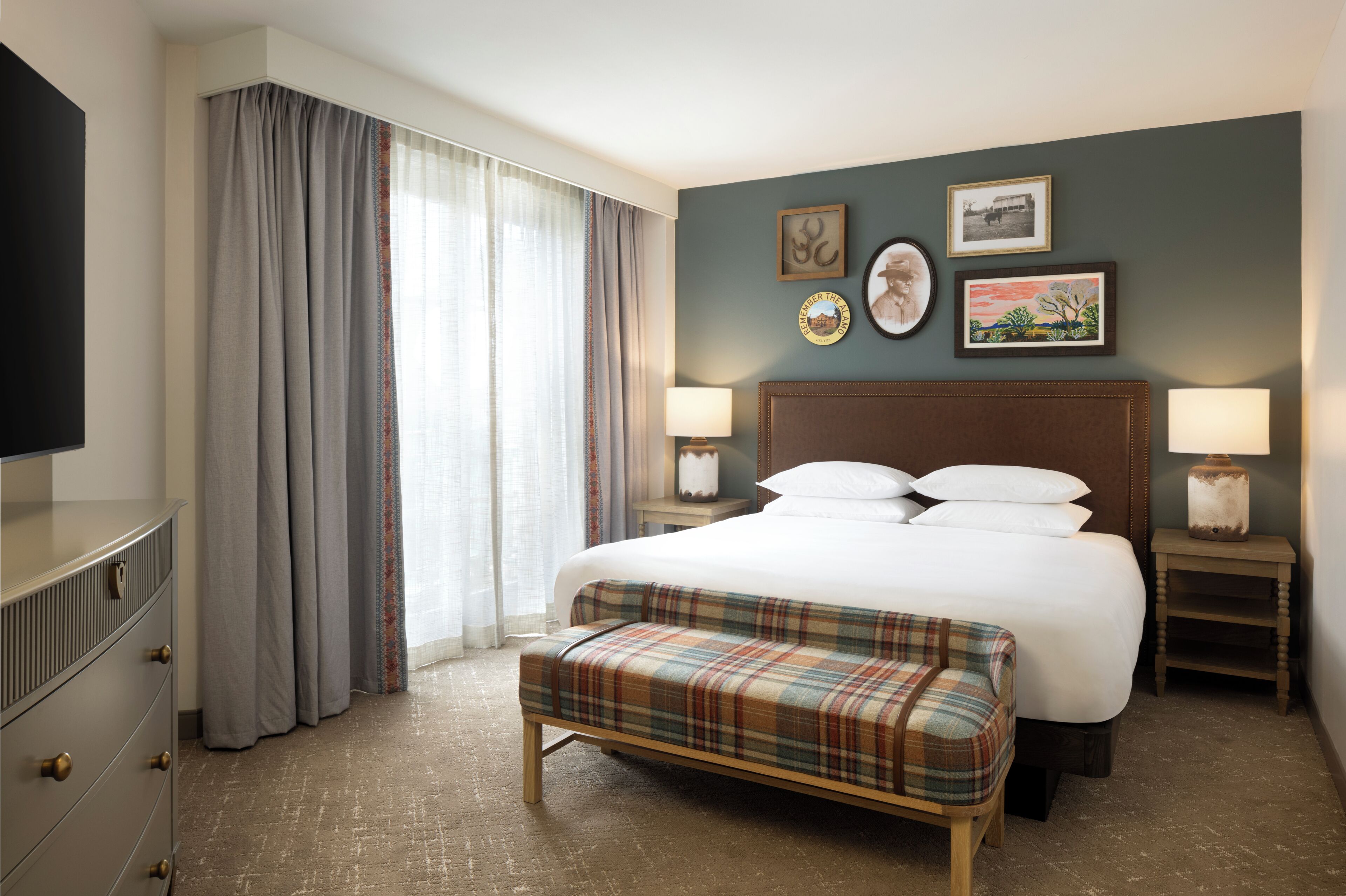 Una habitación de hotel con una cama, dos mesitas de noche, un banco, un televisor y un ventanal grande con cortinas.