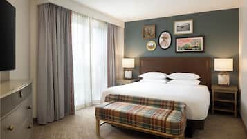 Una habitación de hotel con una cama, dos mesitas de noche, un banco, un televisor y un ventanal grande con cortinas.