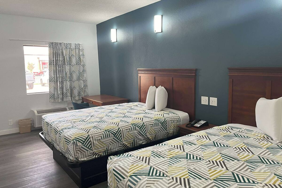 A hotel room with two beds, a small table, a nightstand, and a window with curtains.