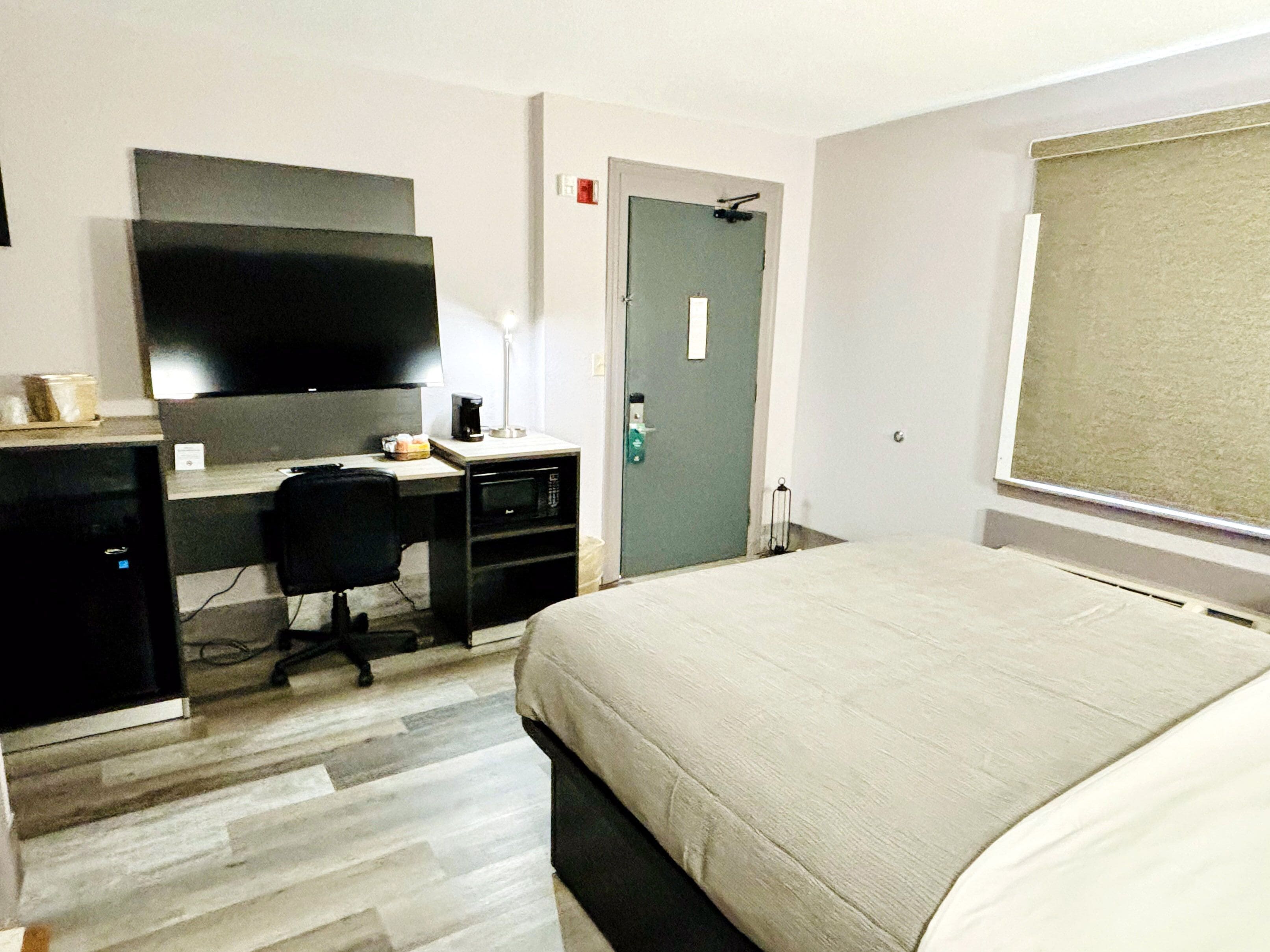 Ein Hotelzimmer mit einem Bett, einem Schreibtisch mit Computer, einem Sessel, einem Fernseher und einer Tür.