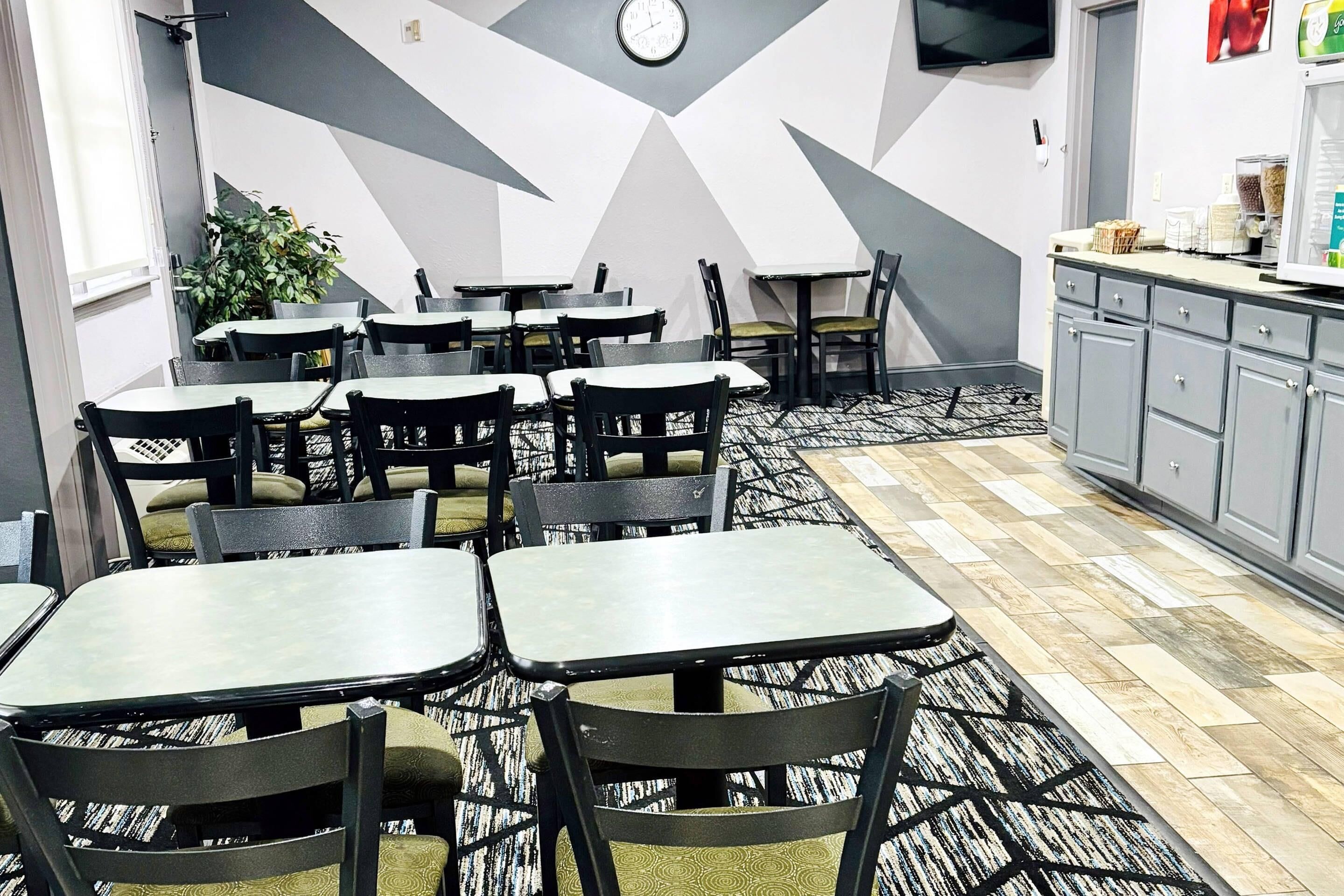 A restaurant interior with tables and chairs, a clock on the wall, and a kitchen area with cabinets and a refrigerator.