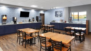 Un restaurant avec des tables et des chaises en bois, un espace cuisine doté d’un mur de crédence bleu, un téléviseur fixé au mur et une fenêtre donnant sur l’extérieur.