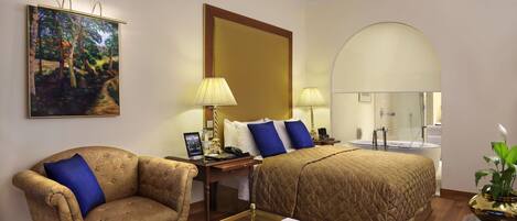 A hotel room with a sofa, a bed, a glass coffee table with apples, a lamp, a painting, and a mirror.