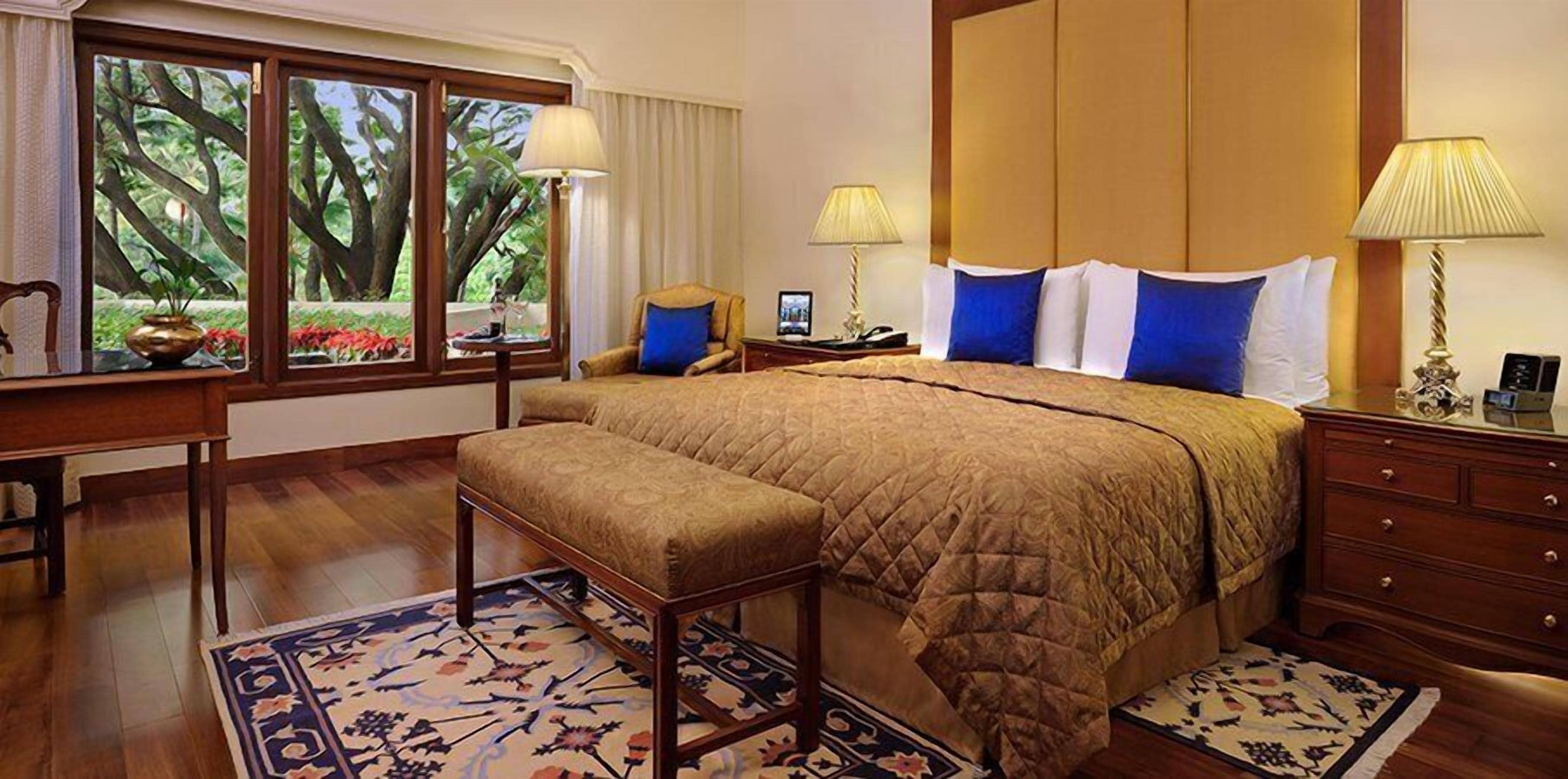 A spacious bedroom with a large bed, a wooden nightstand, a desk, and a chair. There are two windows with curtains, a view of trees, and a patterned rug.