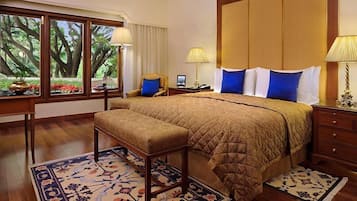 Un dormitorio amplio con una cama grande, una mesita de noche de madera, un escritorio y una silla. Hay dos ventanas con cortinas, vista a árboles y una alfombra estampada.