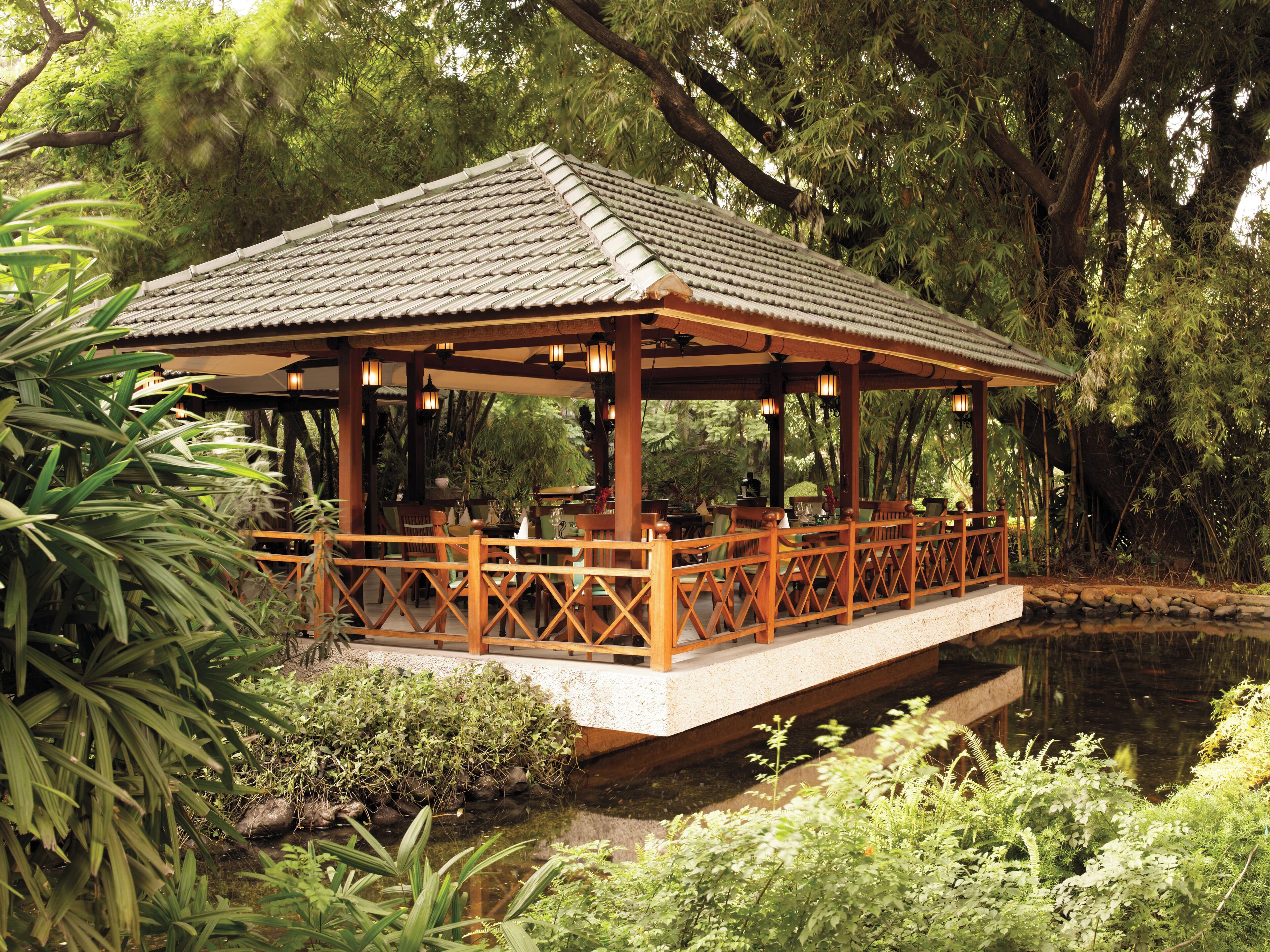 A wooden pavilion with a tiled roof over a pond, surrounded by lush greenery.