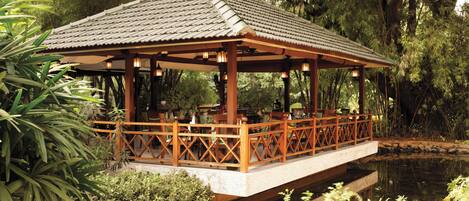 A wooden pavilion with a tiled roof over a pond, surrounded by lush greenery.