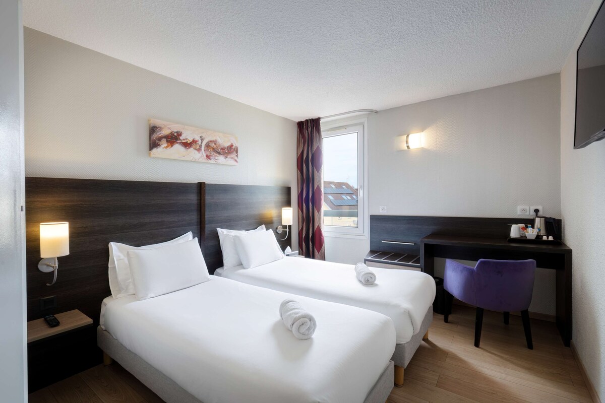 Habitación de hotel con dos camas, un escritorio, una silla, un cuadro y una ventana con cortinas.