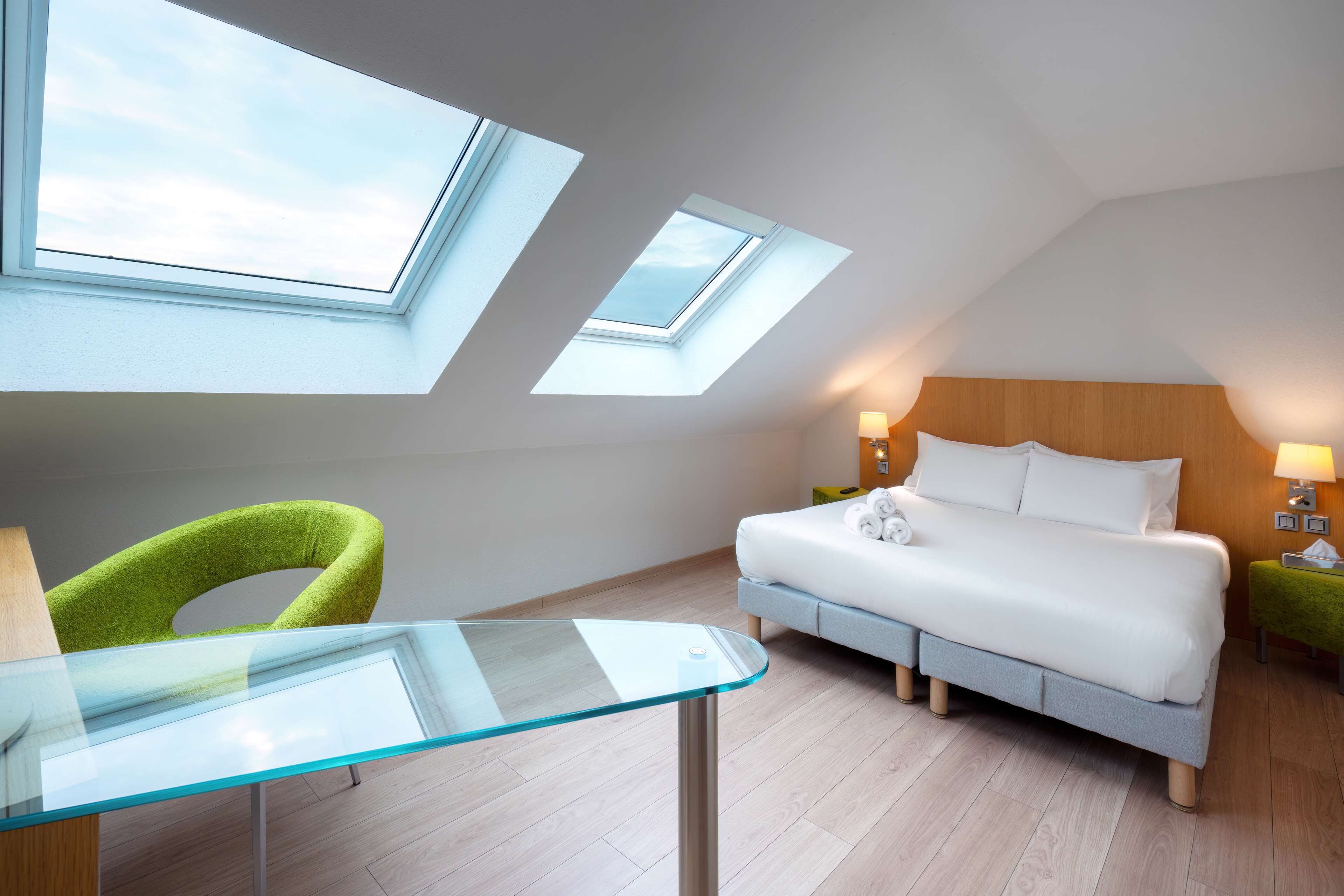 Ein Schlafzimmer mit einem Bett, einem Glastisch, einem grünen Sessel und Dachfenstern.